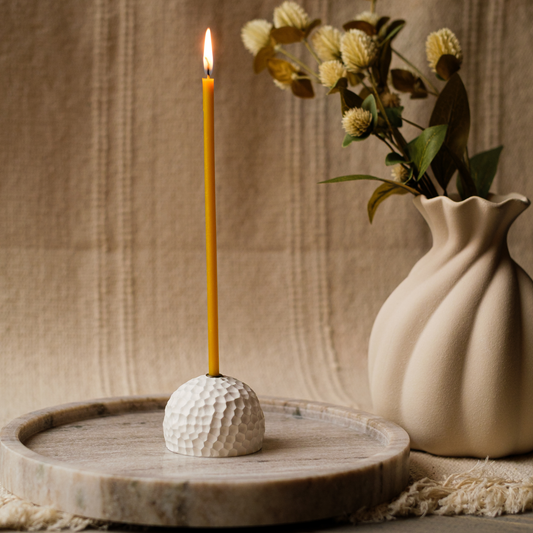 Lit beeswax taper candle in a textured white holder on a round wooden tray, with a white vase of flowers.