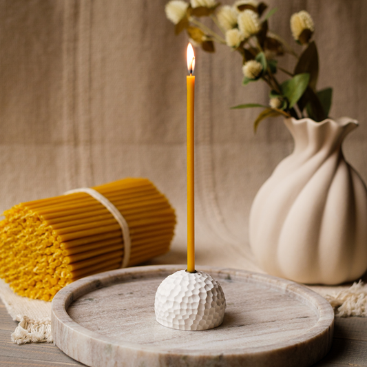 Lit thin beeswax taper candle in a white textured holder on a wooden tray with a vase of flowers and a bundle of tapers in the background.