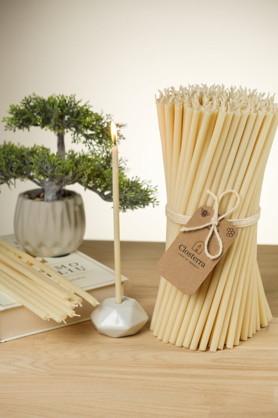 Tall bundle of slim natural beeswax taper candles tied with twine and a kraft Closterra tag, beside a small bonsai and a single taper in a white holder on a light wood table