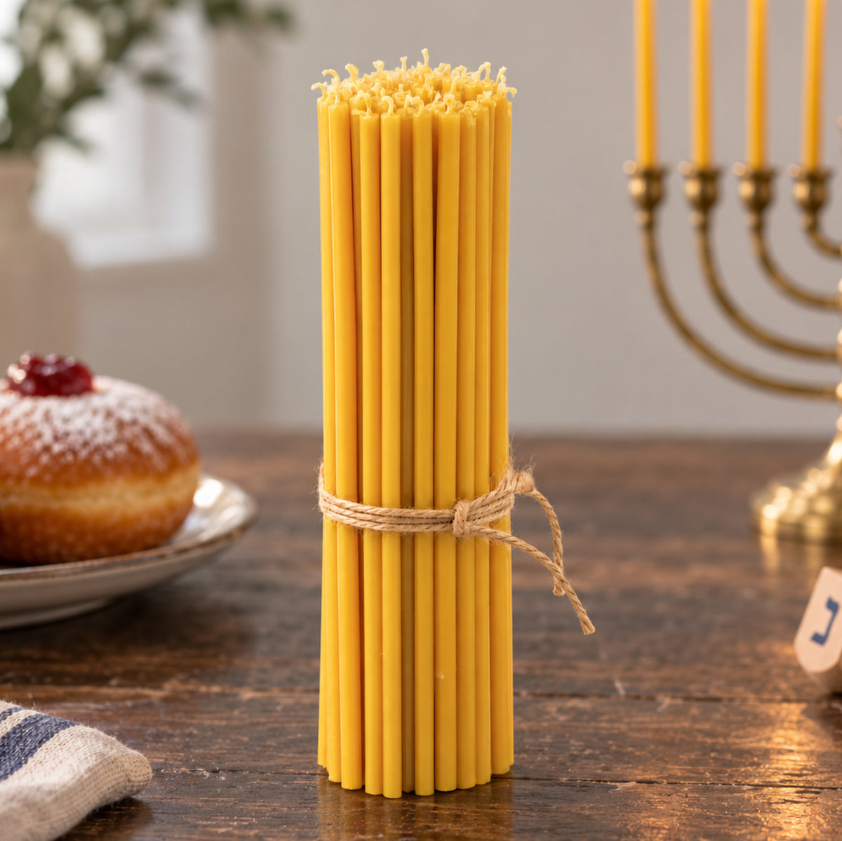 Closterra set of 45 tall 11.6 inch natural yellow beeswax Hanukkah candles tied with twine, beside a brass menorah, a sufganiyah and a dreidel