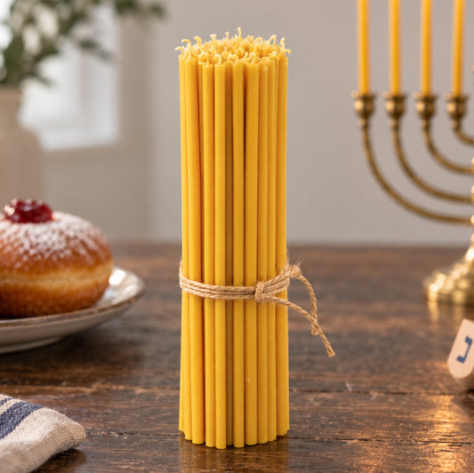 Closterra set of 45 tall 11.6 inch natural yellow beeswax Hanukkah candles tied with twine, beside a brass menorah, a sufganiyah and a dreidel