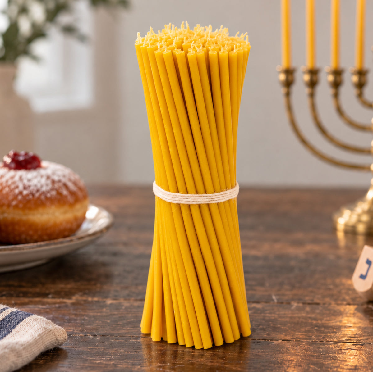 Closterra set of 90 tall 11.6 inch natural yellow beeswax Hanukkah candles tied with string, beside a brass menorah, a sufganiyah and a dreidel
