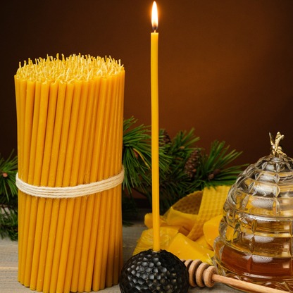 Bundled beeswax taper candles with a lit taper, honey jar, and honey dipper on a rustic surface with greenery.