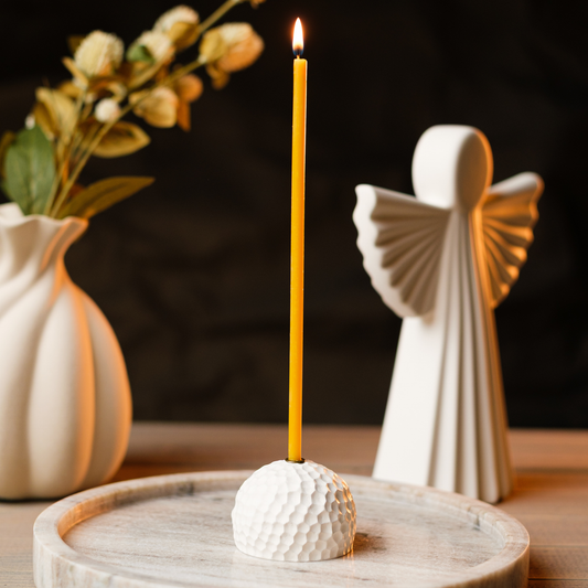 Lit beeswax taper candle in a white holder on a tray, with an angel figurine and flowers in the background.