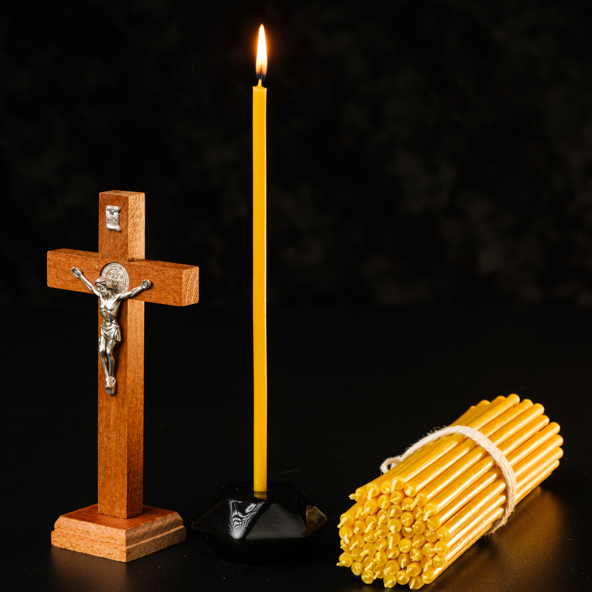 Wooden cross beside a lit beeswax taper candle and a bundle of beeswax tapers on a black background.
