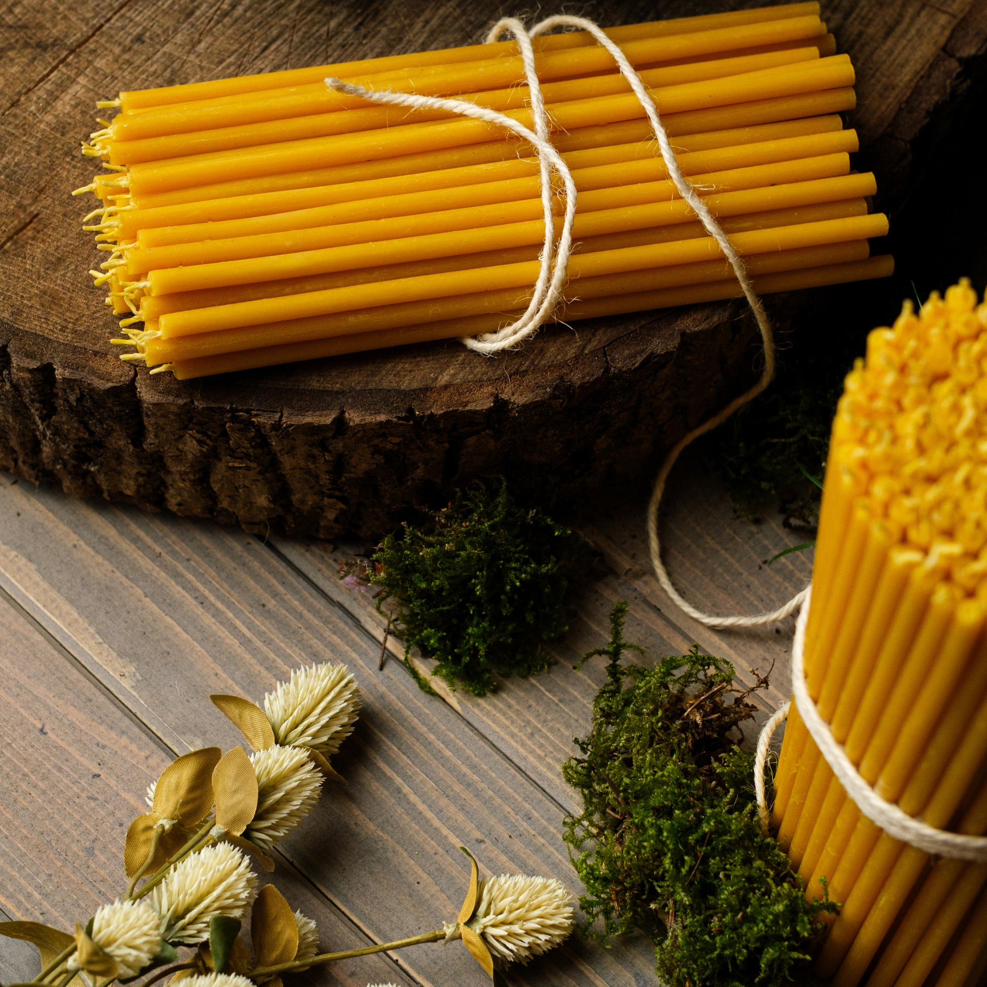 Bundles of thin beeswax taper candles tied with twine on a rustic wooden surface with moss and greenery.
