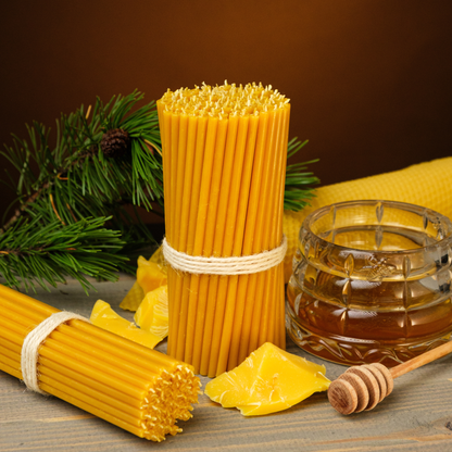Two bundles of thin beeswax taper candles with beeswax pieces, a honey jar, and honey dipper on a wooden surface with pine greenery.