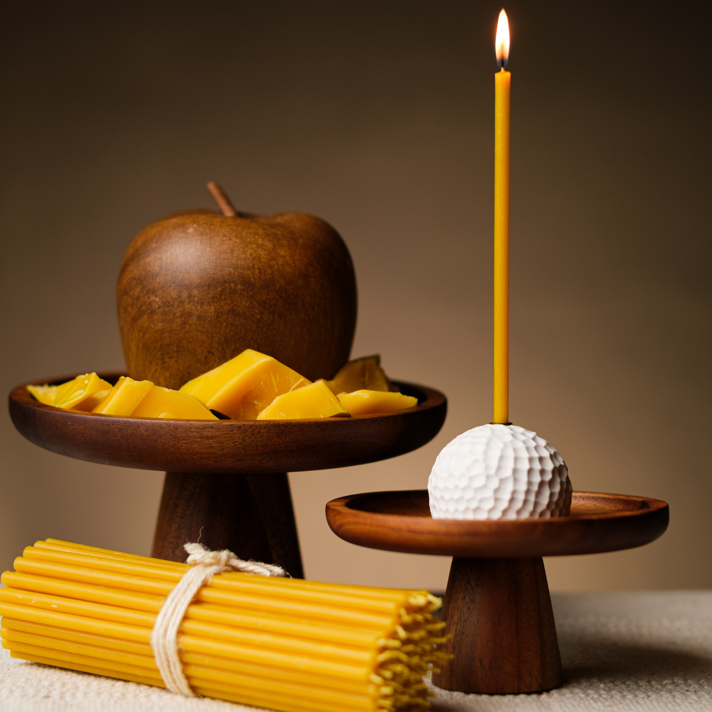 Lit beeswax taper candle in a white holder beside bundled thin beeswax tapers and beeswax pieces in a warm still life.


