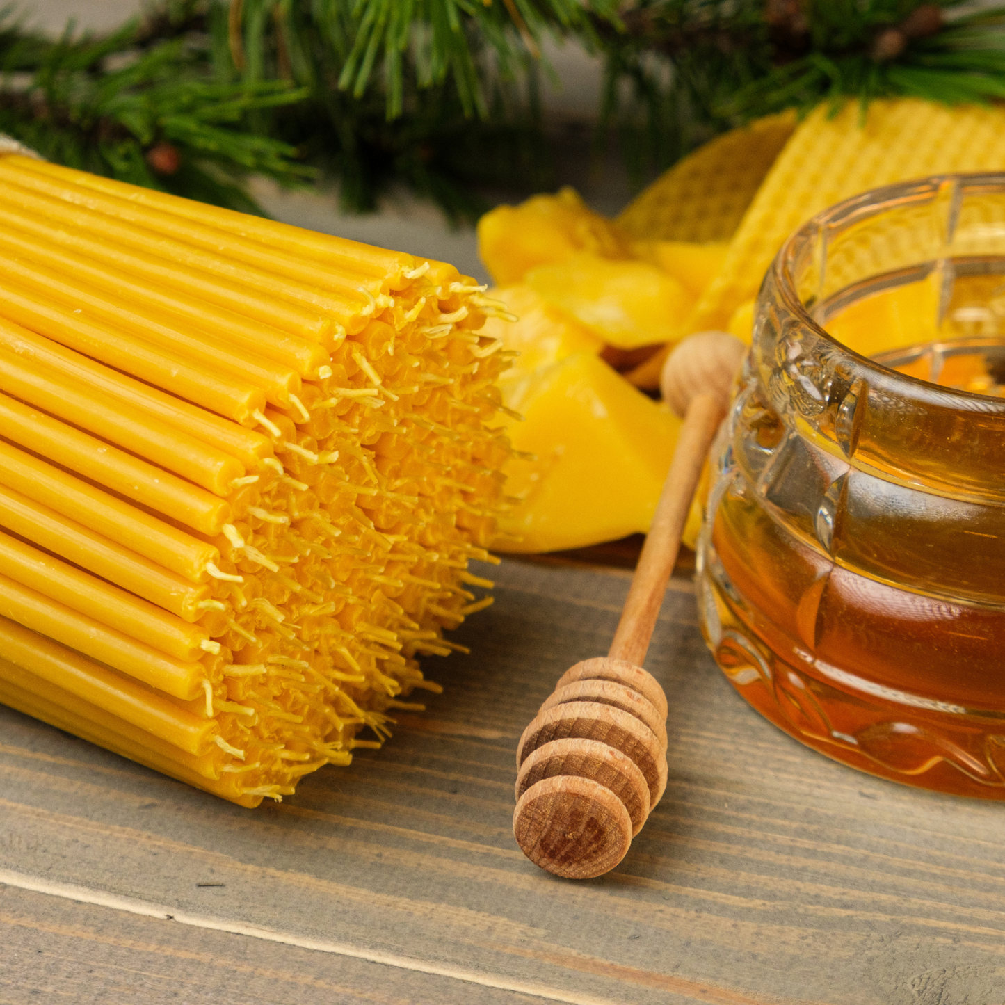 Close-up of a bundle of thin beeswax taper candles beside a honey jar and wooden honey dipper on a rustic surface with evergreen greenery.