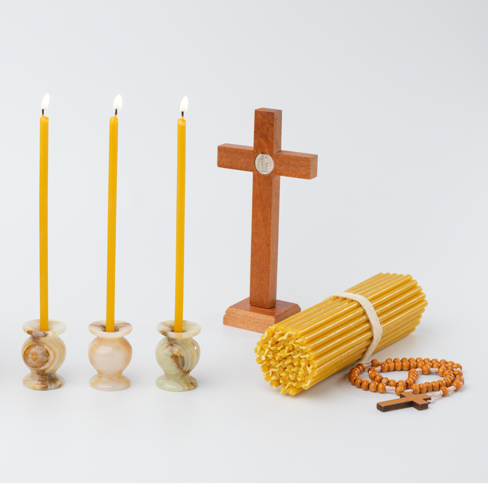 Three lit candles in marble holders, a wooden cross, a bundle of yellow candles, and a rosary on a white background.