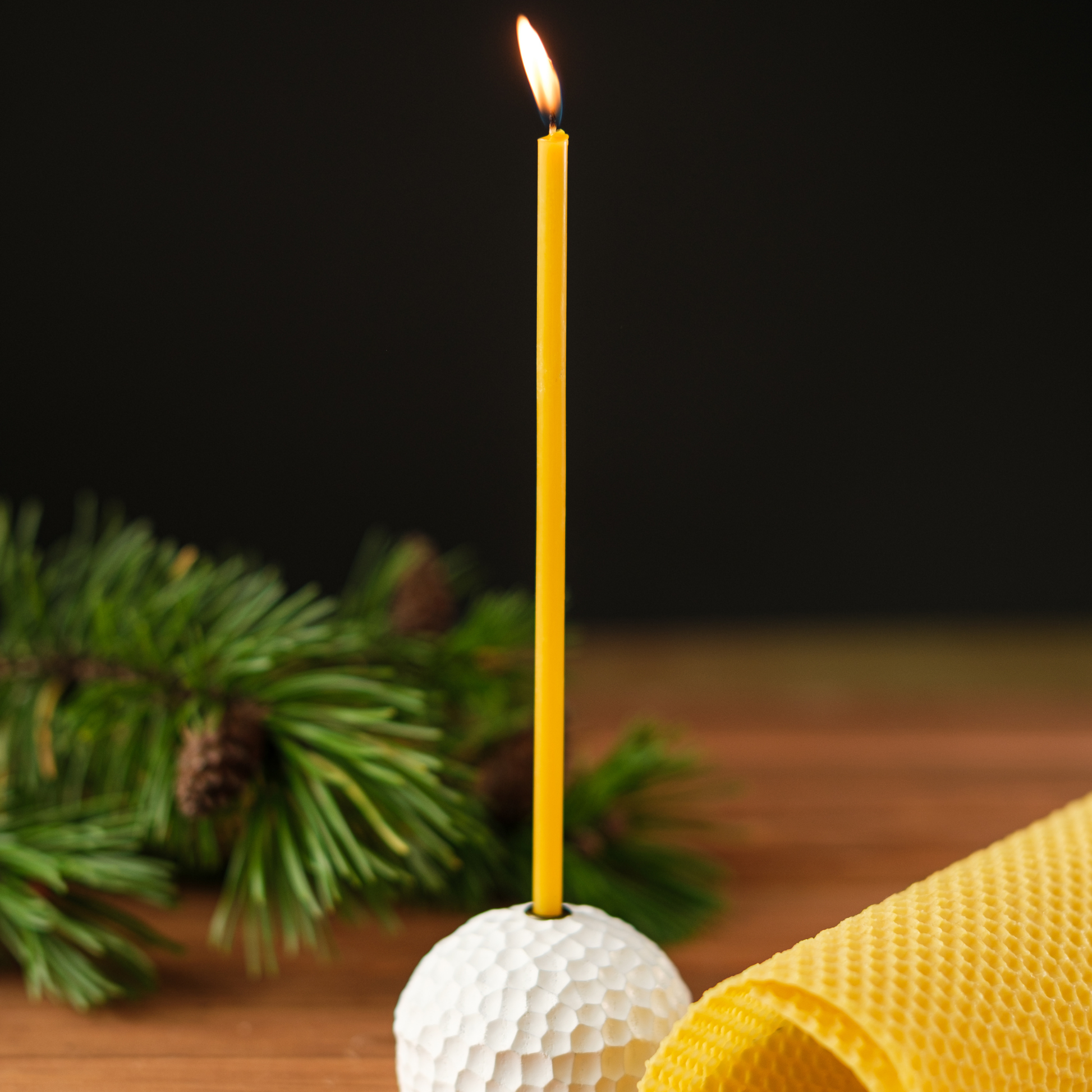 Lit thin beeswax taper candle in a white textured holder on a wooden surface with evergreen greenery and a rolled beeswax sheet.