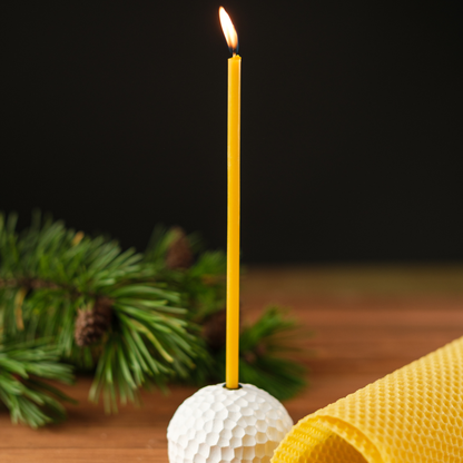 Lit thin beeswax taper candle in a white textured holder on a wooden surface with evergreen greenery and a rolled beeswax sheet.