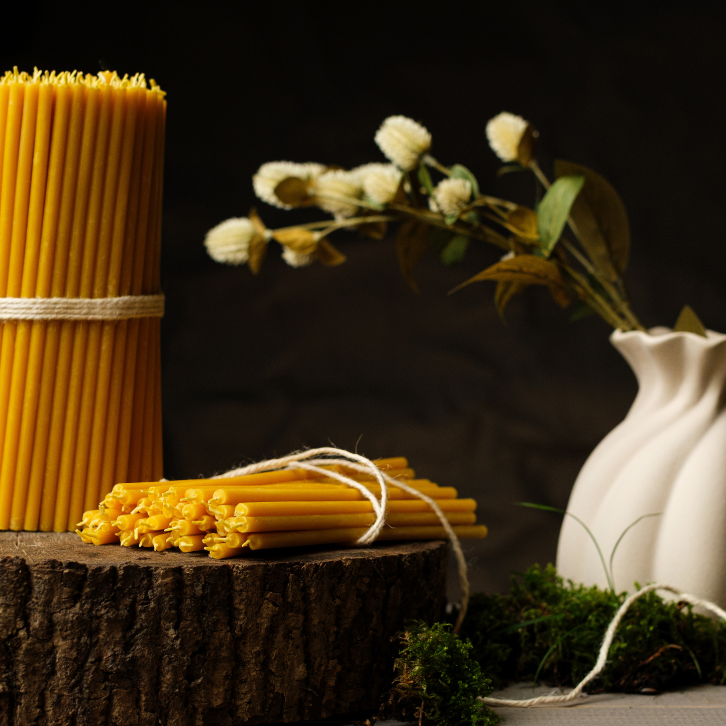 Large bundle of thin beeswax taper candles beside a smaller tied bundle on a wood slice with a white vase of flowers on a dark background.