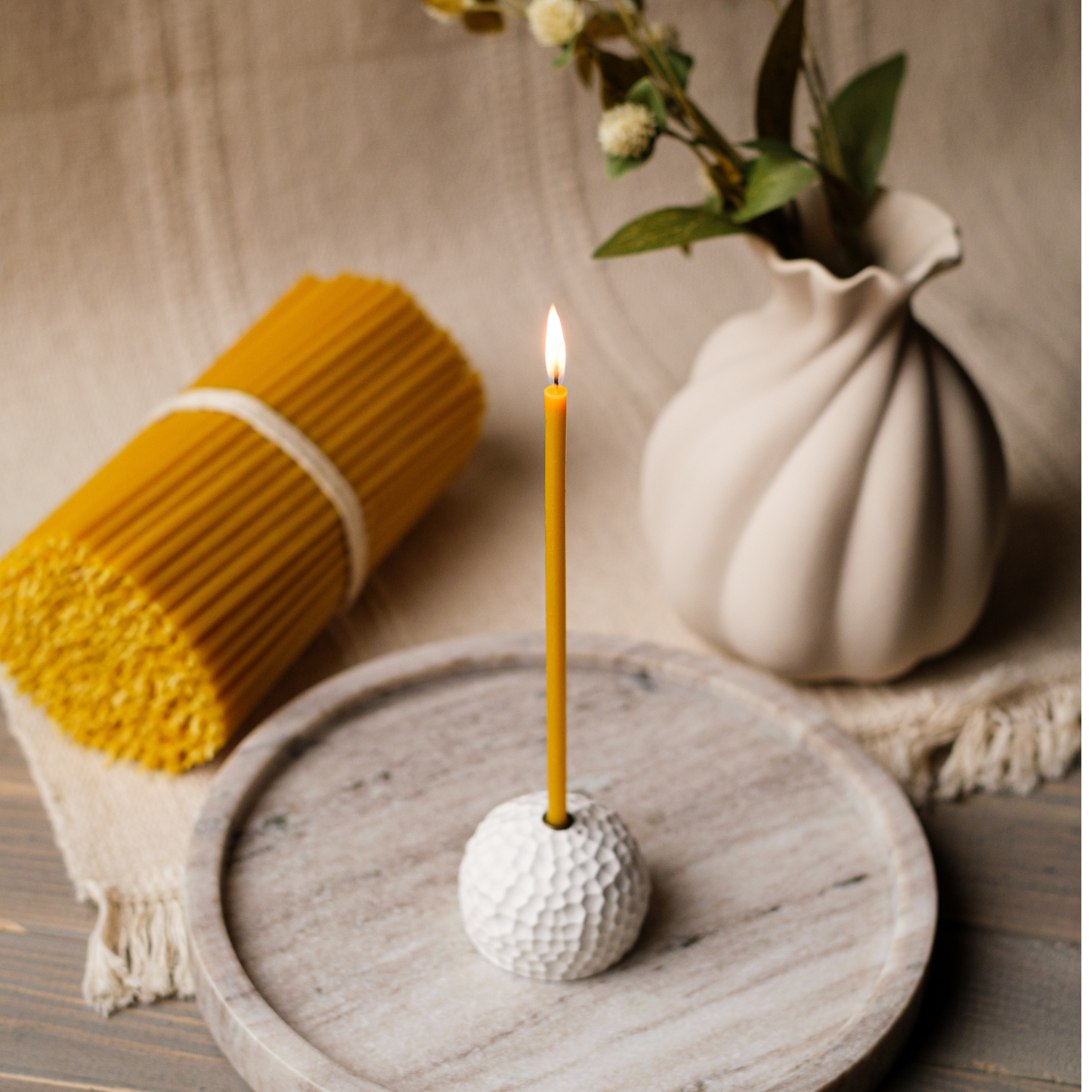 “Pure beeswax taper candle in a white golf-ball holder on a wooden tray beside a bundle of thin tapers and a white vase.”