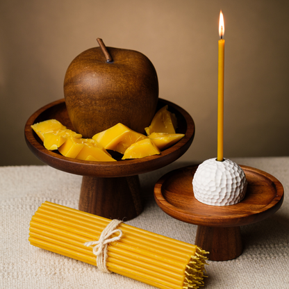 Lit beeswax taper candle in a white textured holder on a wooden stand, with bundled tapers and beeswax pieces.