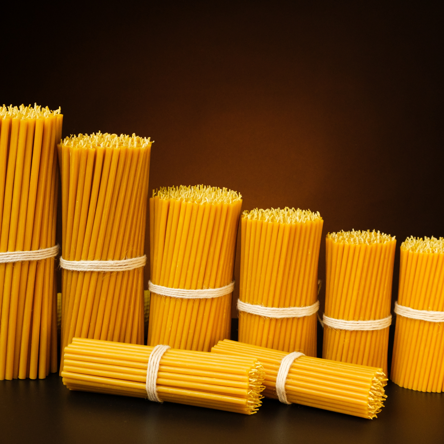 “Assorted bundles of thin pure beeswax taper candles arranged by size on a dark background.”