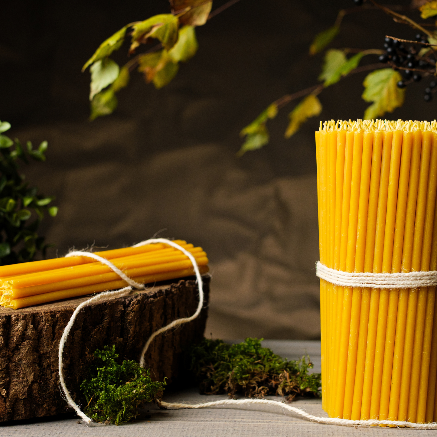 Two bundles of thin beeswax taper candles tied with twine on a rustic wooden surface with greenery in the background.

