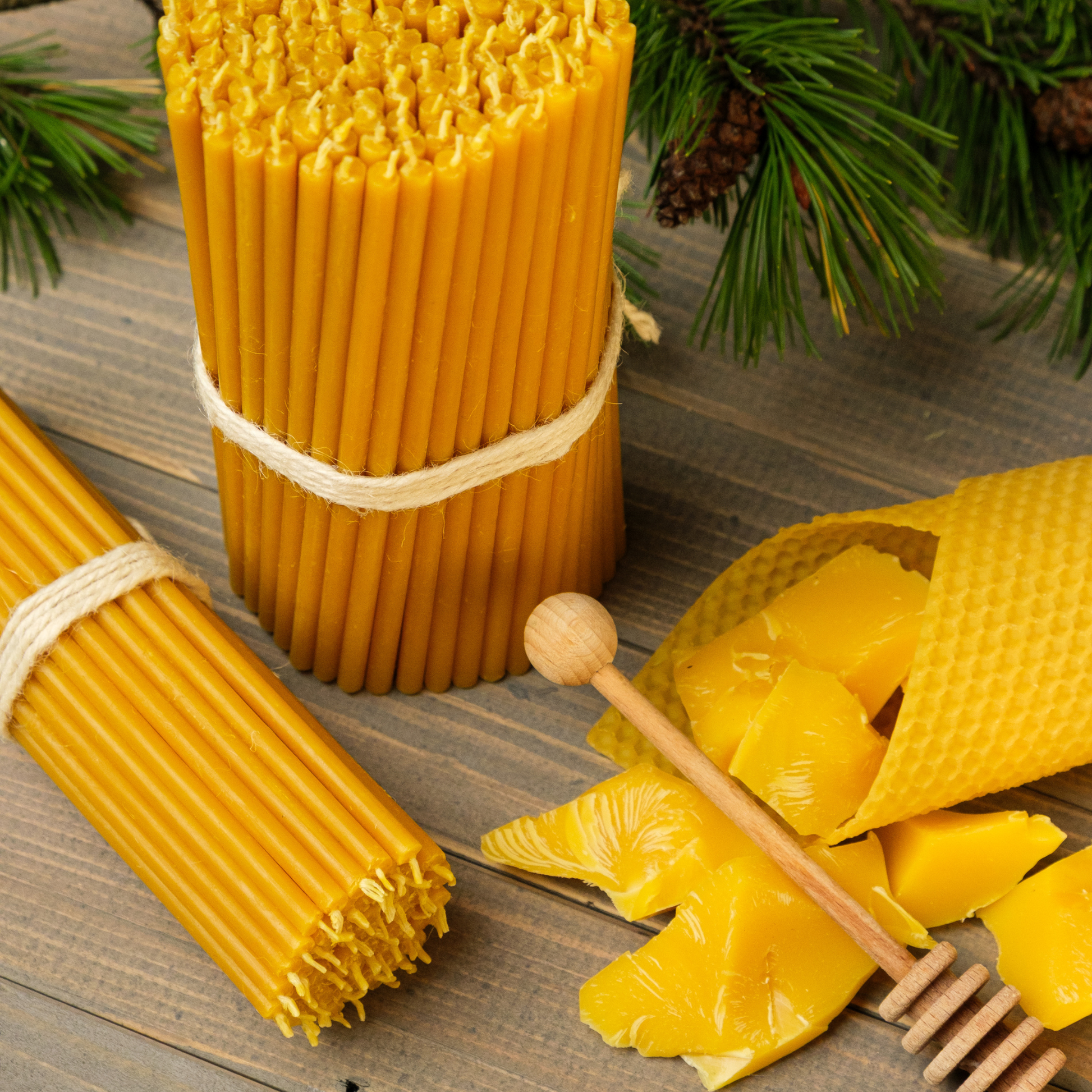 Bundles of thin beeswax taper candles with beeswax blocks and a honey dipper on a wooden surface with greenery.