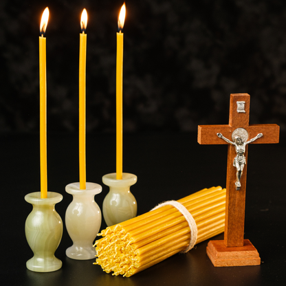 Three lit beeswax taper candles in holders with a wooden crucifix and a bundle of thin beeswax tapers on a dark background.