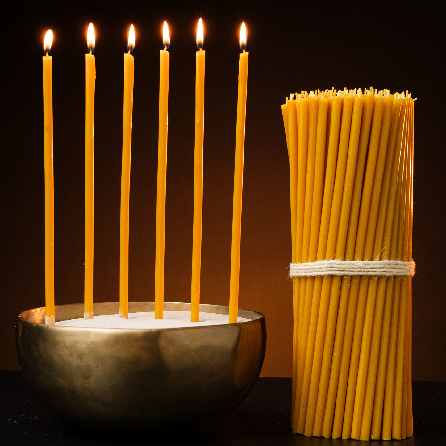Six lit thin beeswax taper candles in a bowl beside a large bundle of beeswax tapers tied with twine on a dark background.