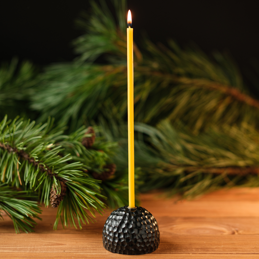 Lit thin beeswax taper candle in a dark textured holder on a wooden surface with evergreen branches in the background.