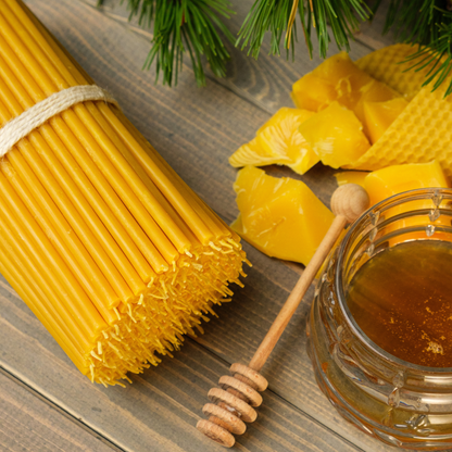 Bundle of thin beeswax taper candles with a honey jar, honey dipper, and beeswax pieces on a wooden surface.