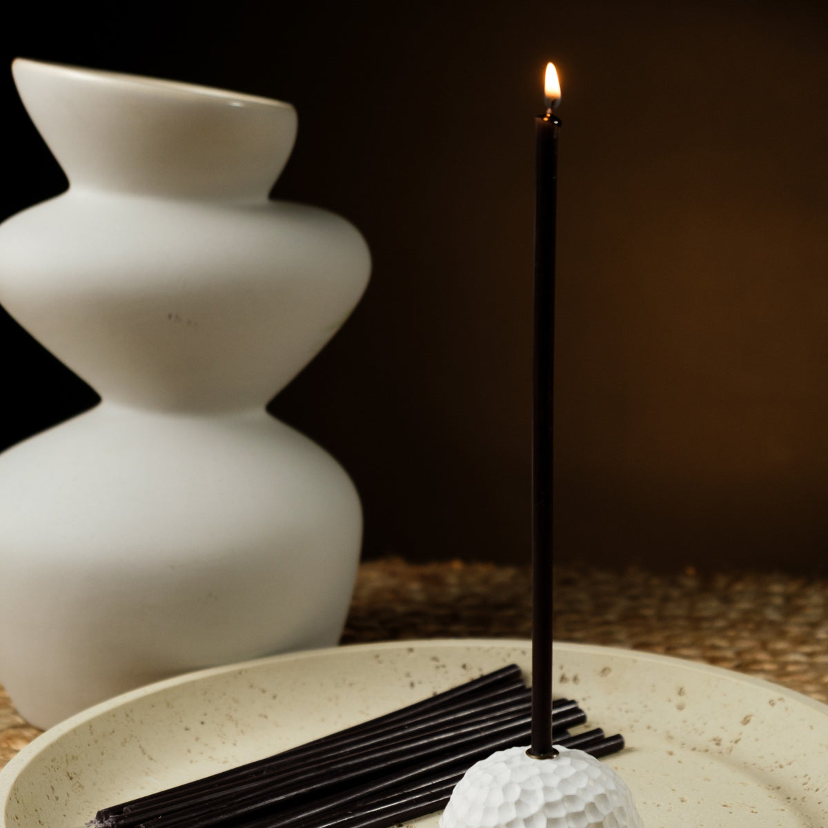 Black beeswax taper candles 6.3″, lit taper in a white holder beside a white vase