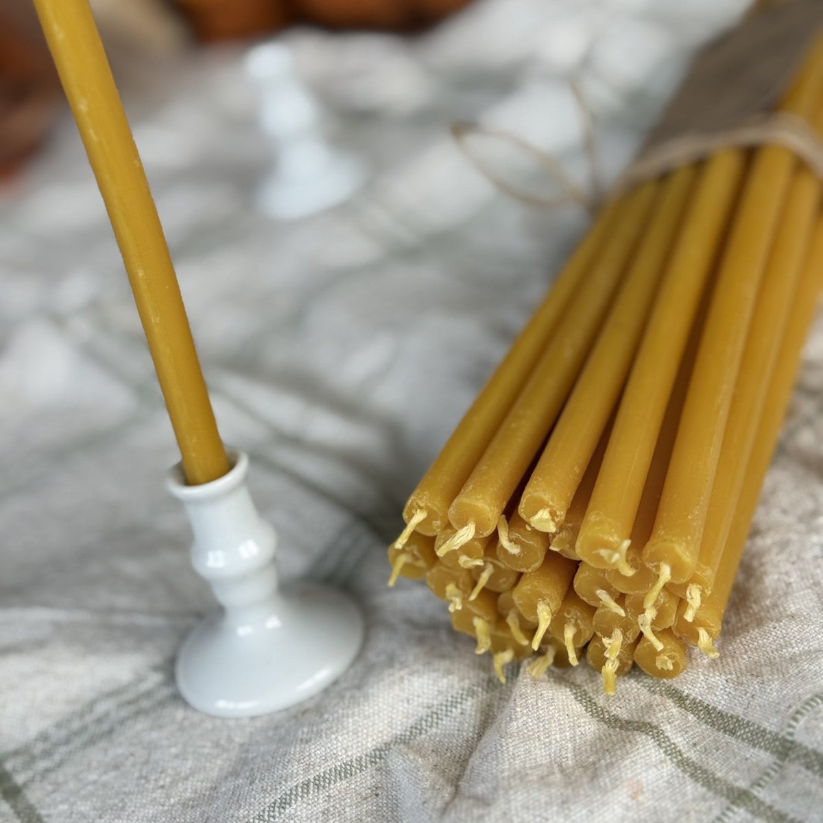 Natural yellow beeswax taper candles 11.6″, bundle of 25 beside a taper in a white candlestick