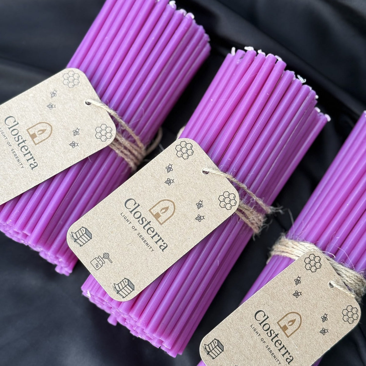 Purple beeswax taper candles, row of tagged bundles on black satin