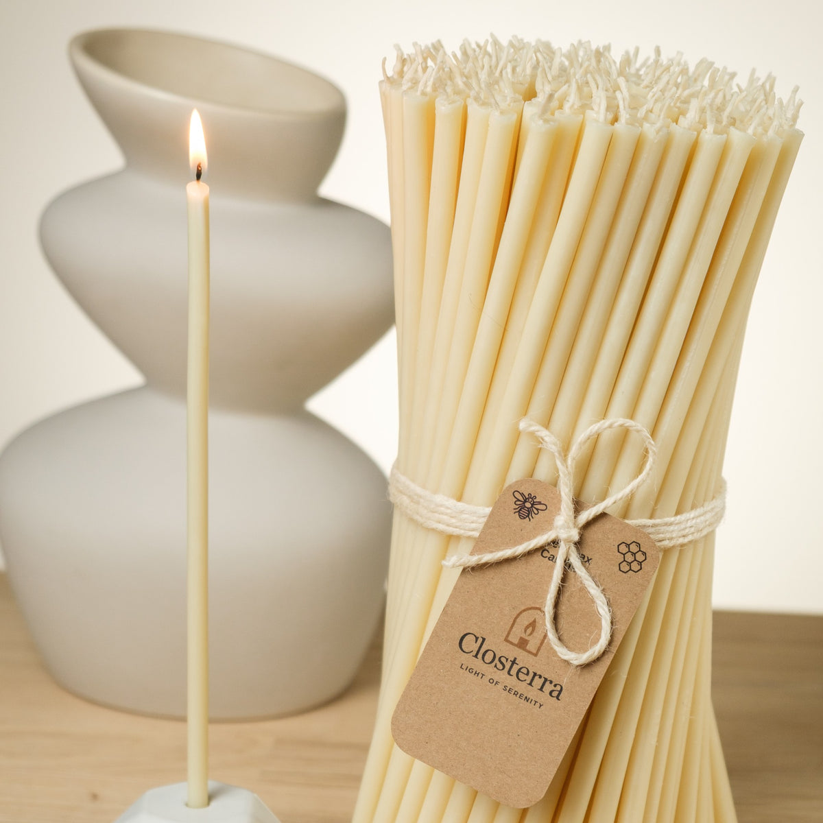 White beeswax taper candles 10.4″, lit taper beside a bundle with a Closterra tag and a white vase